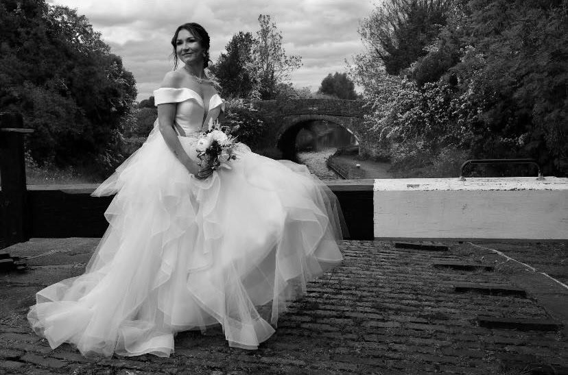 Bride in wedding dress holding bouquet near water with lush greenery.